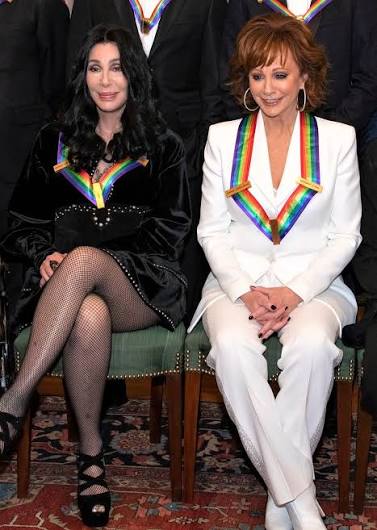 Cher and Reba McEntire seated together, both wearing rainbow honoree sashes